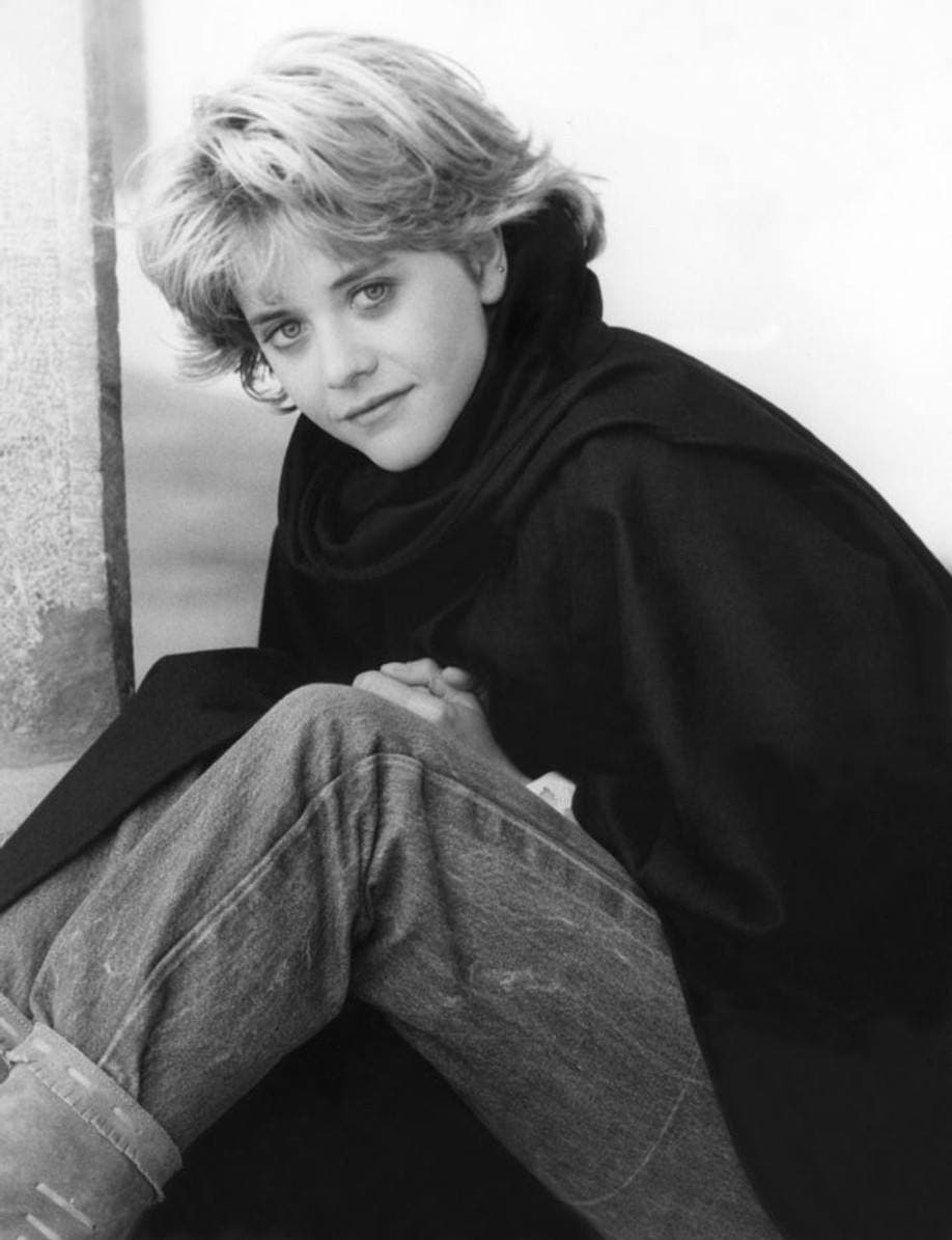 Meg Ryan with short fair hair, sitting with her knees up in a dark coat, in black and white