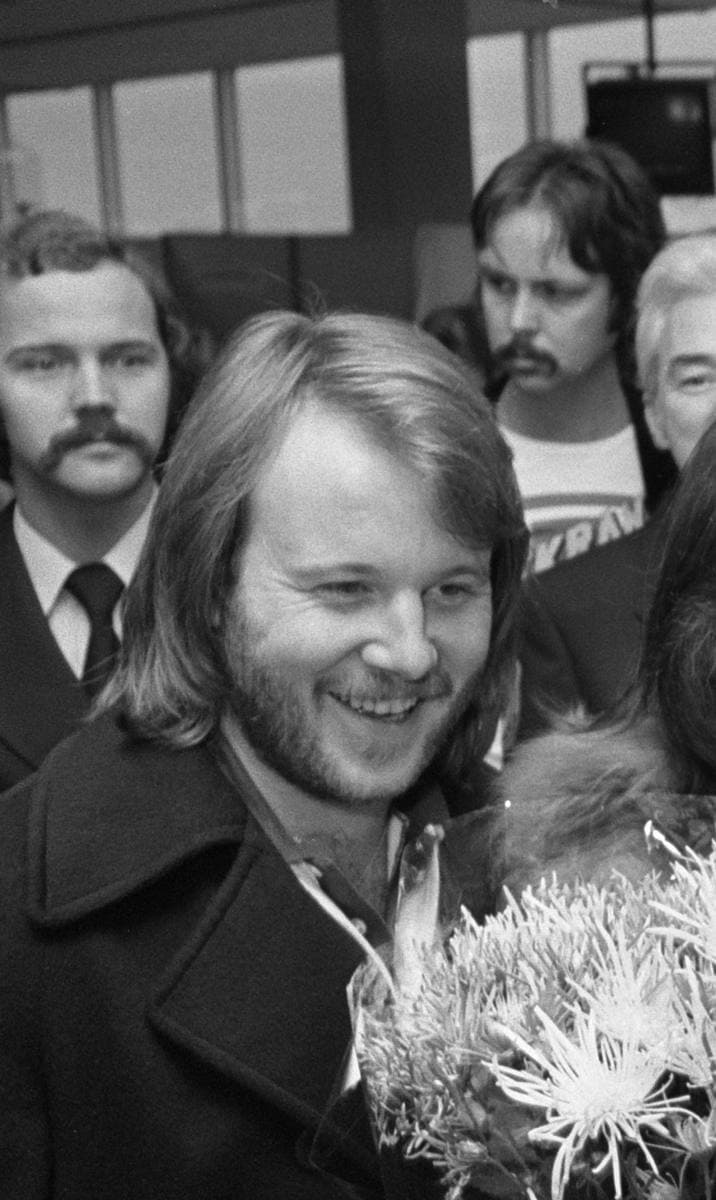 Benny Andersson, bearded and smiling, in a dark coat in an airport crowd
