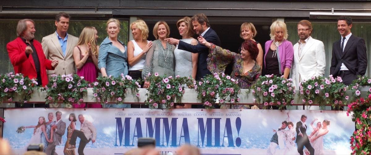 A row of people waving from a flower-lined balcony above a large Mamma Mia! banner