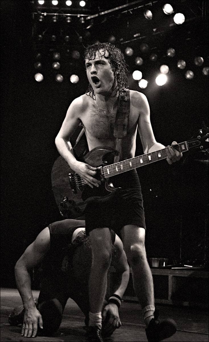 Angus Young, shirtless in school shorts, playing a guitar under stage lights