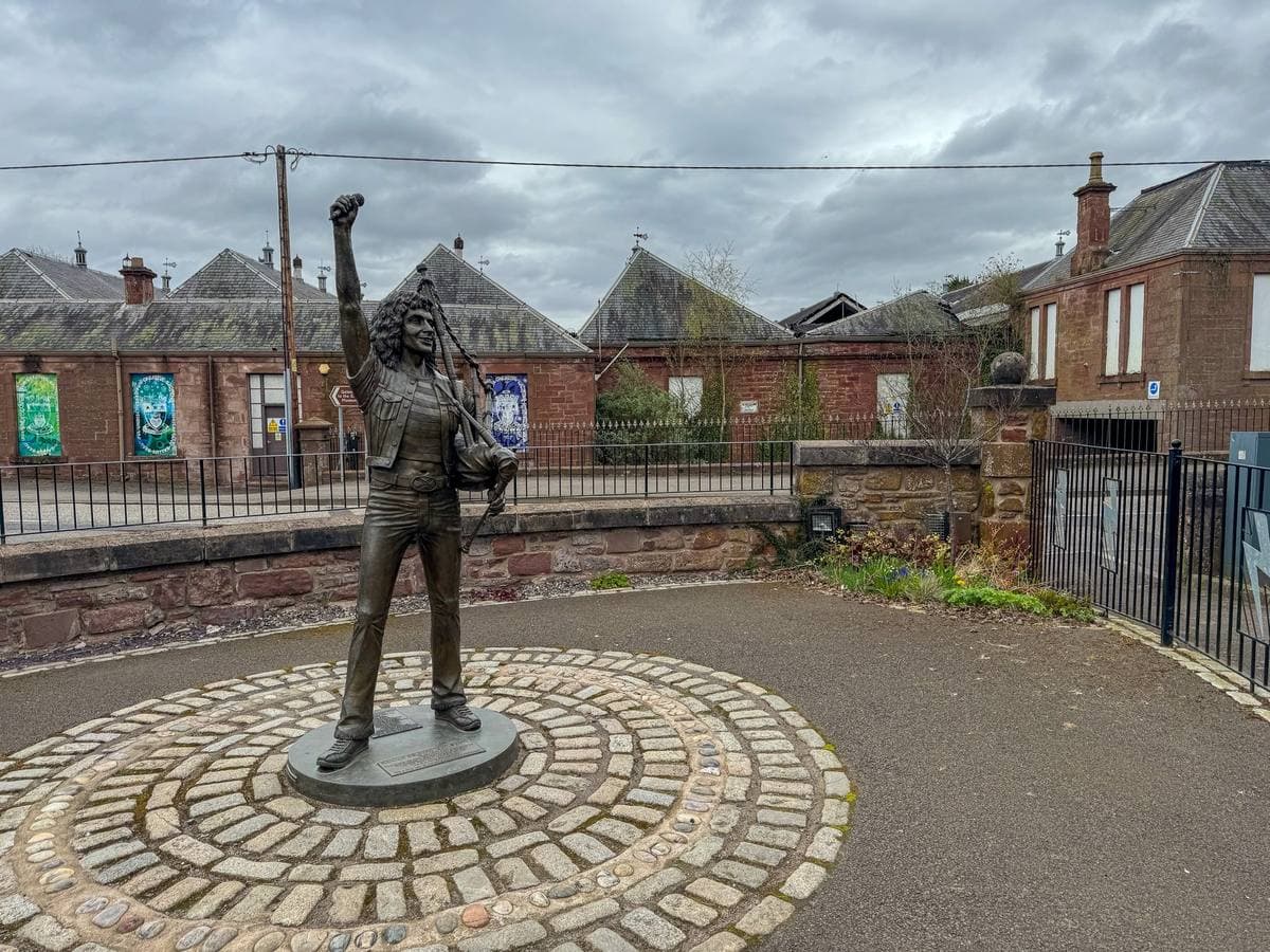 A bronze statue with one arm raised, standing on a round mosaic in a small garden