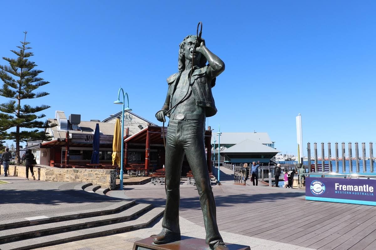 A bronze statue of a singer with a microphone raised, on a plinth by a harbour