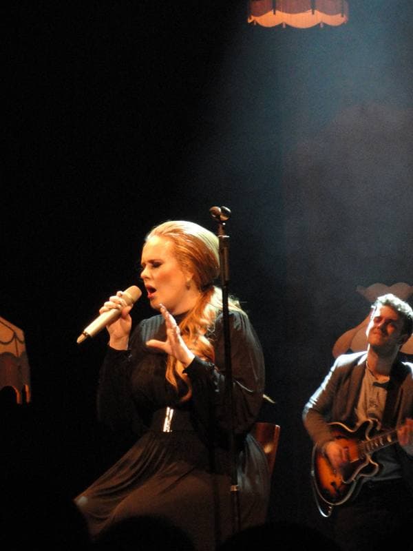 Adele singing on stage in a black dress under warm light