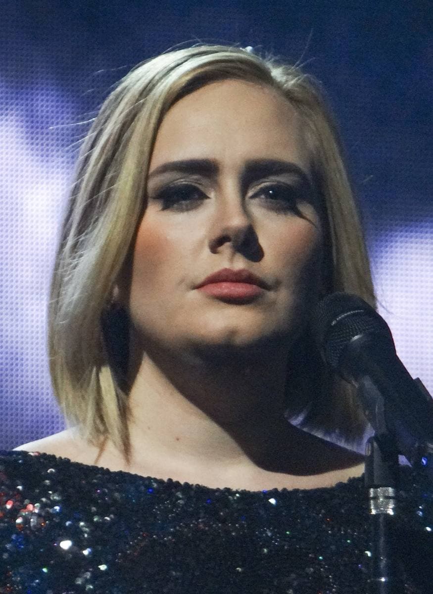 Adele in close-up, blonde hair to her shoulders and dark winged eyeliner, in a sequinned black dress beside a microphone