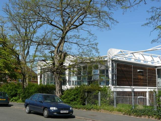 A low modern school building with a curved glass front behind trees on a suburban road