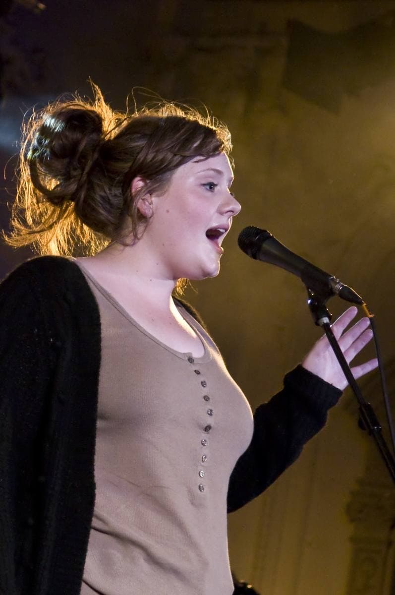 A young Adele, hair pinned up, singing at a microphone in a wood-panelled hall