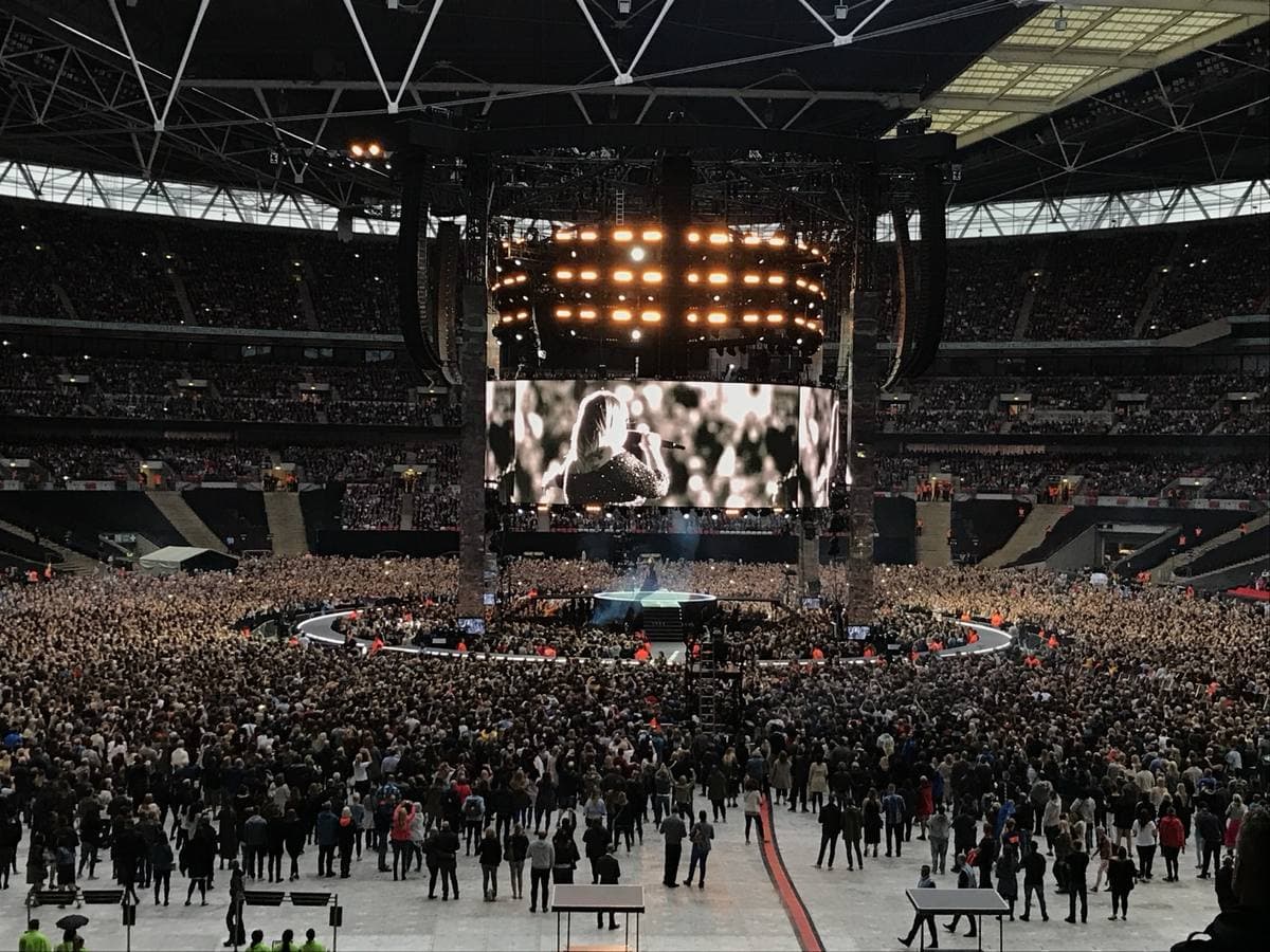 A full Wembley Stadium in daylight, a giant screen hung over a stage in the middle of the pitch
