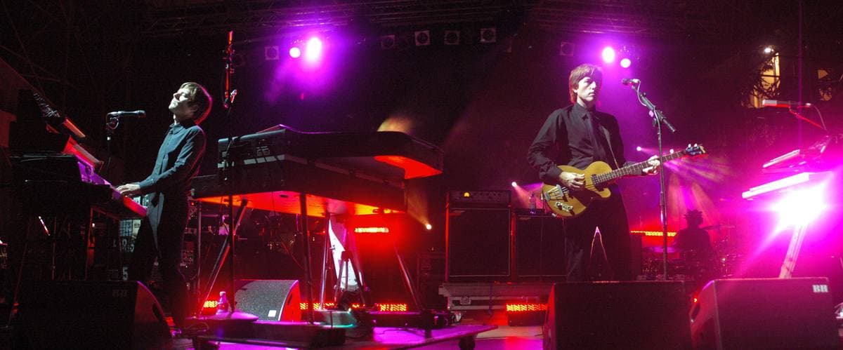 Jean-Benoît Dunckel at a keyboard on the left and Nicolas Godin with a bass guitar on the right, both in black, under magenta stage light