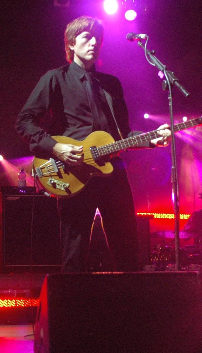 Nicolas Godin in a black shirt and tie playing a pale bass guitar at a microphone