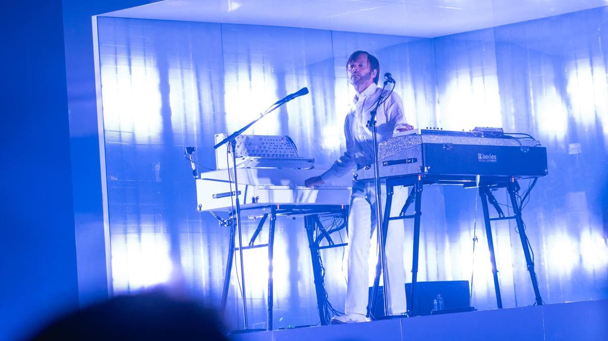 A musician in white at a stack of keyboards inside a box of blue light