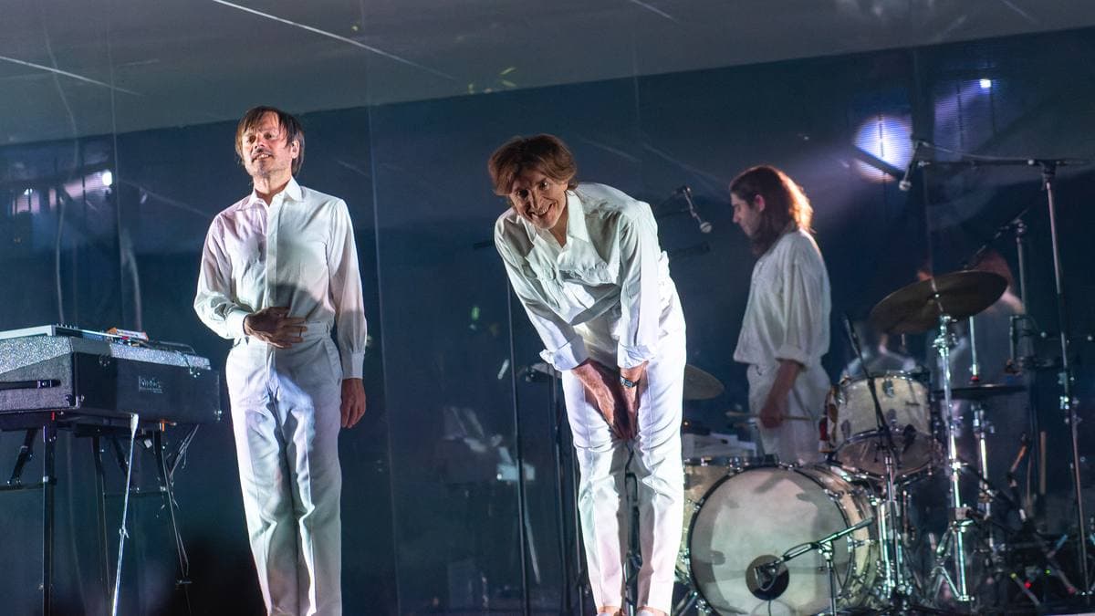 Two men in white suits bowing on a white-lit stage beside a drum kit