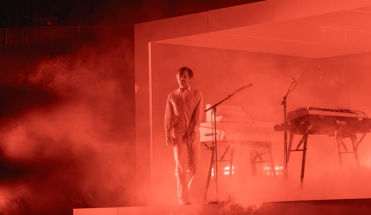 A musician in white standing inside a glowing red, smoke-filled box on a dark stage with keyboards