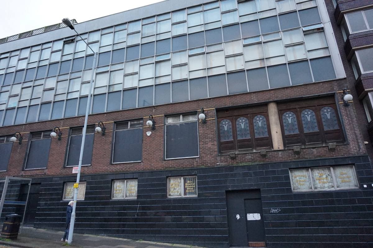 A long building with a dark tiled ground floor and rows of windows above, beside a sloping pavement