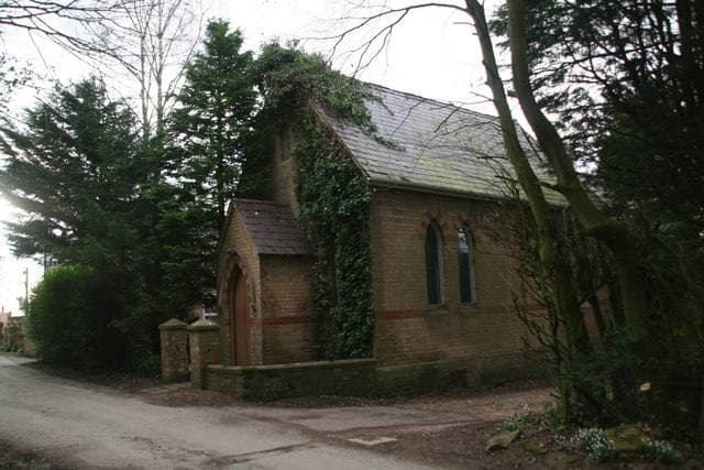 A small brick chapel with a pitched roof half hidden by ivy and trees beside a lane