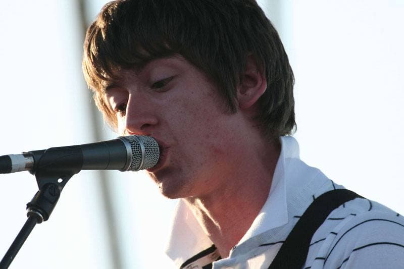 Close-up of Alex Turner singing into a microphone in daylight, his fringe over his eyes and a striped collar at his neck