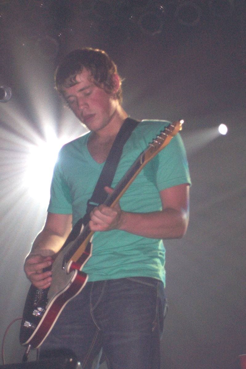 Jamie Cook in a green T-shirt playing a red guitar, a stage light flaring behind his shoulder