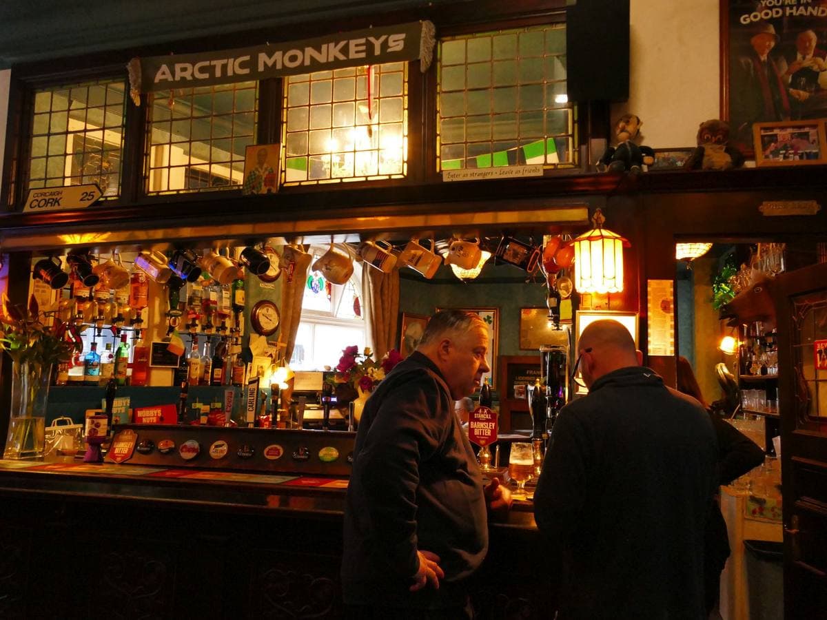 A warmly lit pub bar with two drinkers, a sign reading ARCTIC MONKEYS above the bar back