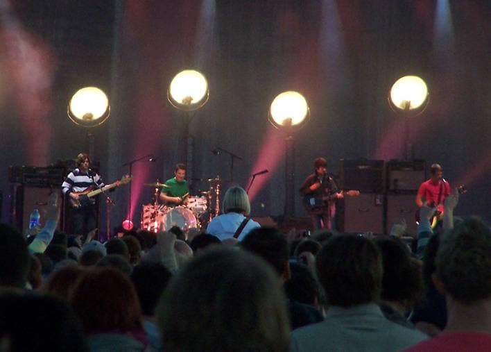 Four large globe lights glowing above the band on stage, pink light behind them, heads of the crowd in front