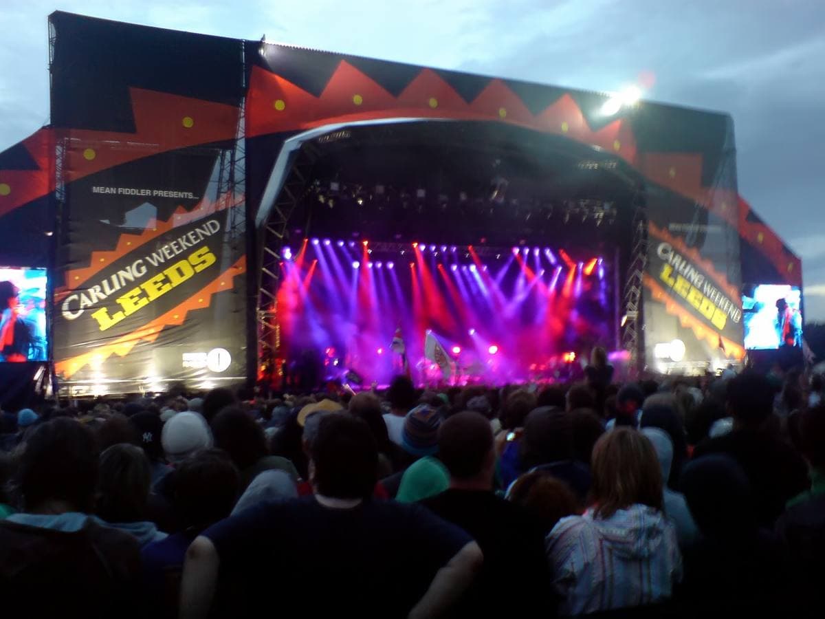 A festival main stage at dusk lit pink and purple, CARLING WEEKEND LEEDS on the side screens, a crowd in the foreground