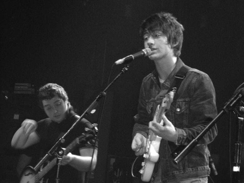 Black and white: Andy Nicholson bent over his bass on the left, Alex Turner in a denim jacket singing into the microphone on the right