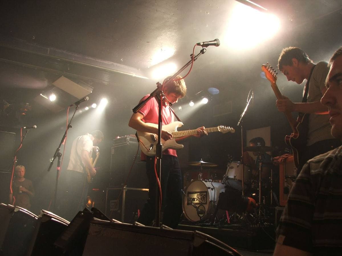 The band on a small club stage under white lights, two guitarists at the front and the drum kit behind them