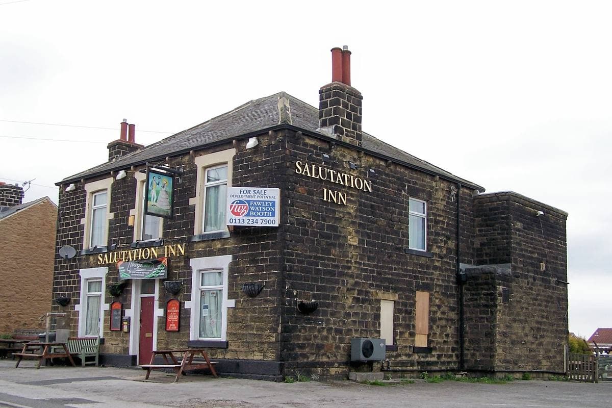 A stone-built corner pub with a hanging sign and benches out front