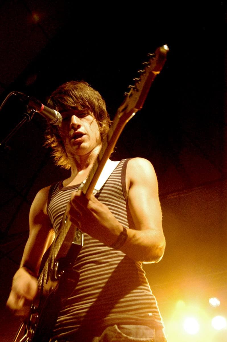 Alex Turner with a mop of dark hair and a striped vest, singing at the microphone with his guitar raised, lit gold from below