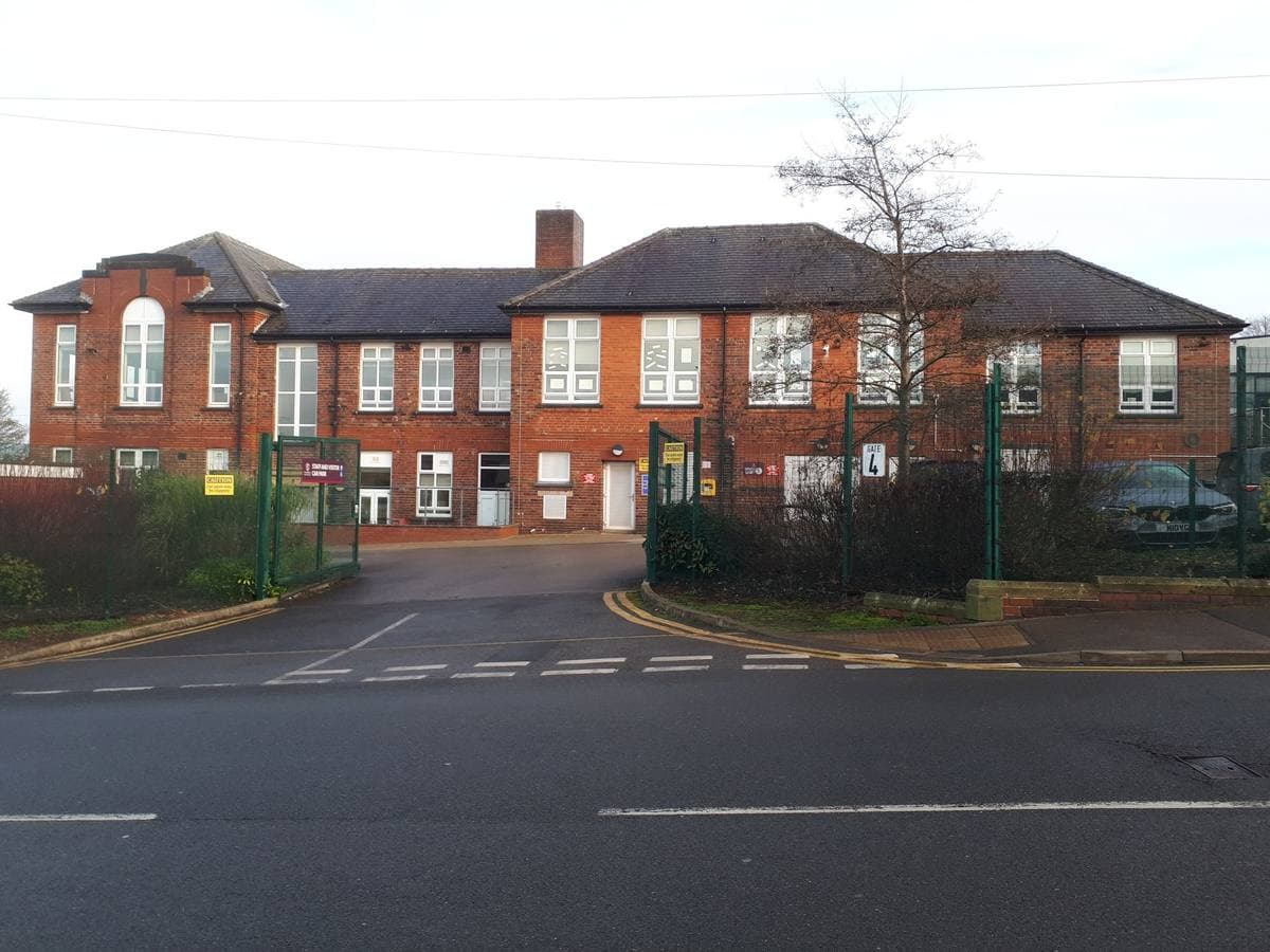 Red-brick school buildings with tall windows behind a gated entrance and a quiet road