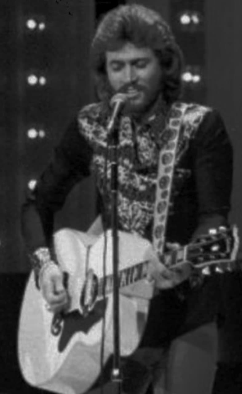 Barry Gibb singing with a guitar on a lit television stage