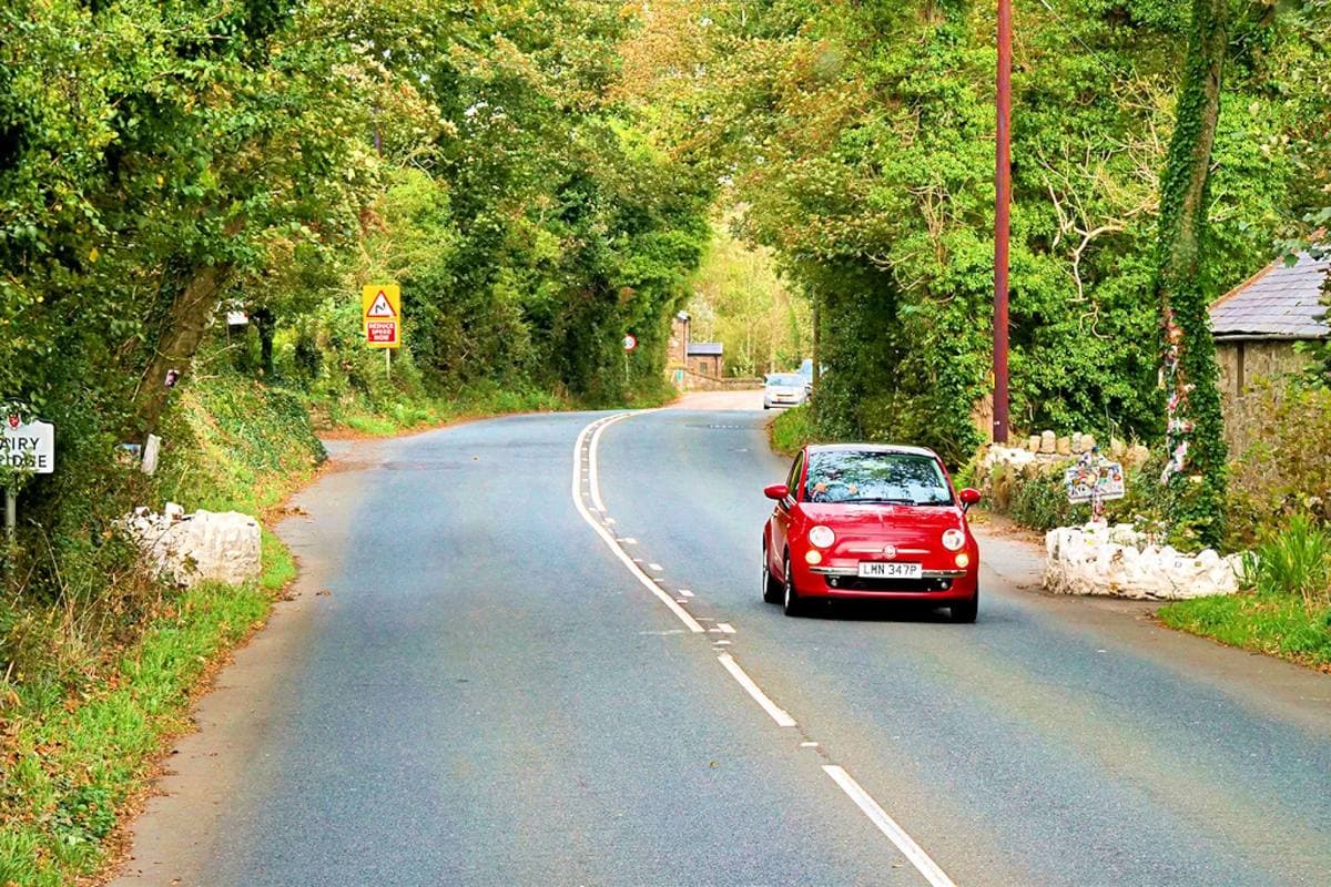 A small red car crossing a low bridge on a tree-lined country road