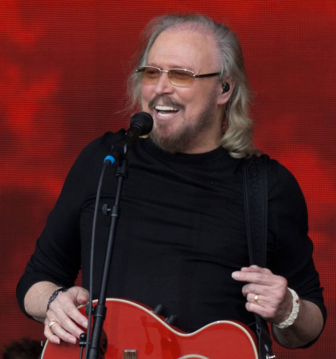 Barry Gibb, white-haired and smiling in tinted glasses, singing with a red guitar