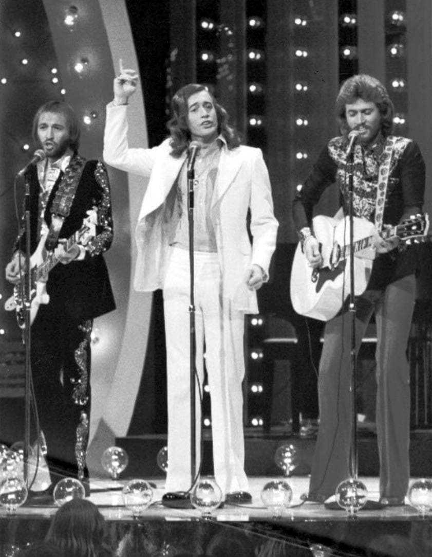 Three young men with long hair singing at microphones on a television stage lined with lights, one in a white suit between two with guitars