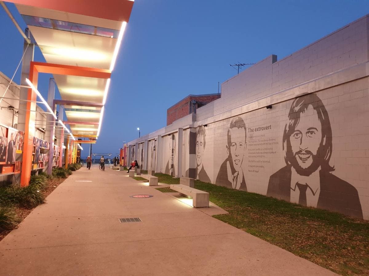A lit walkway at dusk beside a long grey wall painted with large portraits of the brothers