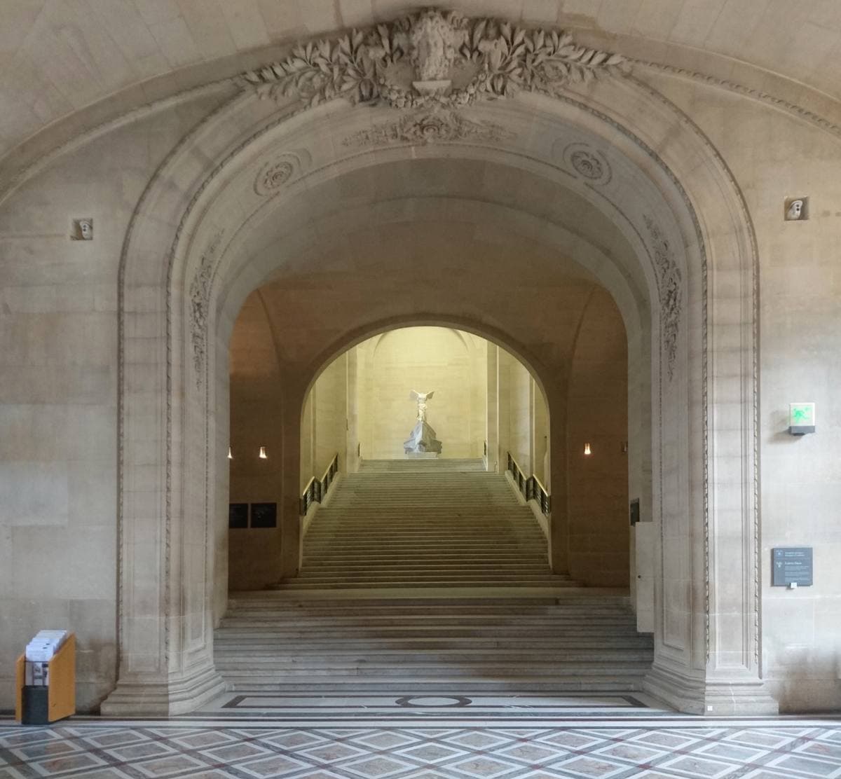 A broad stone staircase under an arch rising to a white winged statue at the top