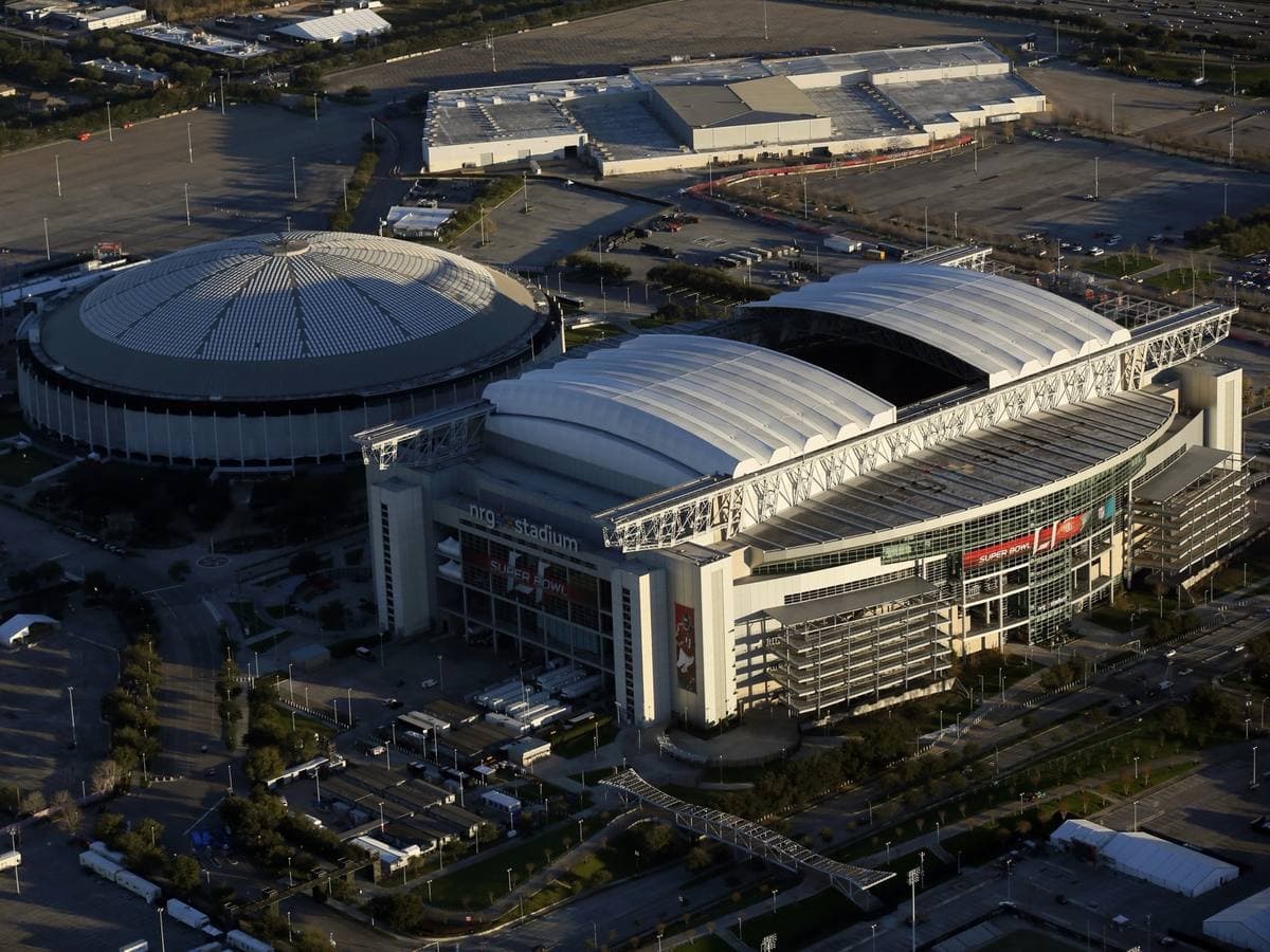 An aerial view of a large stadium with a white roof next to an older round domed stadium