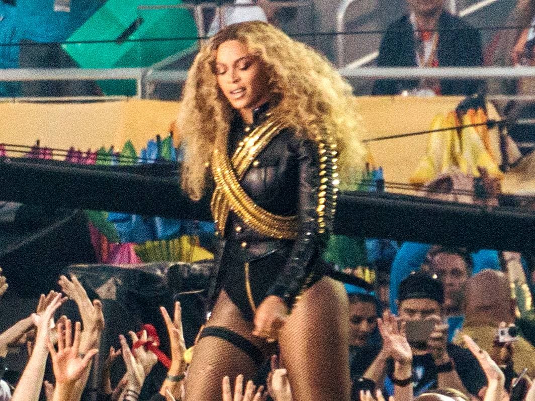 A woman in a black jacket with gold bandoliers and big blonde curls stands above a crowd of raised hands