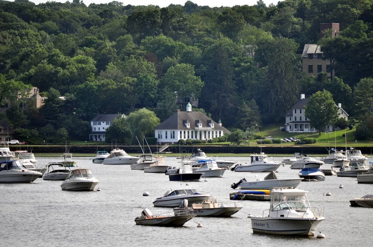 Small motorboats moored in a calm harbour below wooded hills and white houses