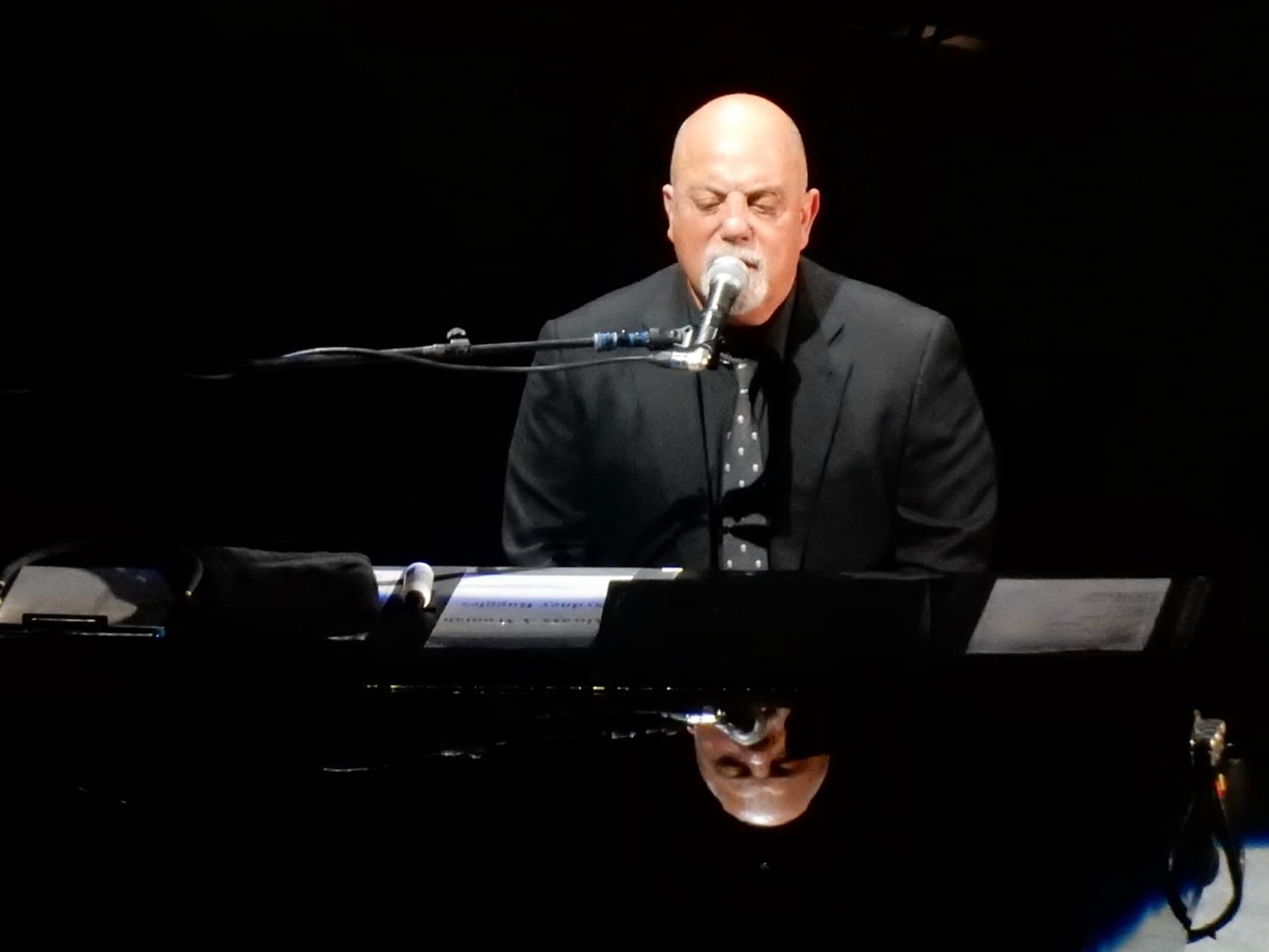 Billy Joel singing at a black piano on a dark stage