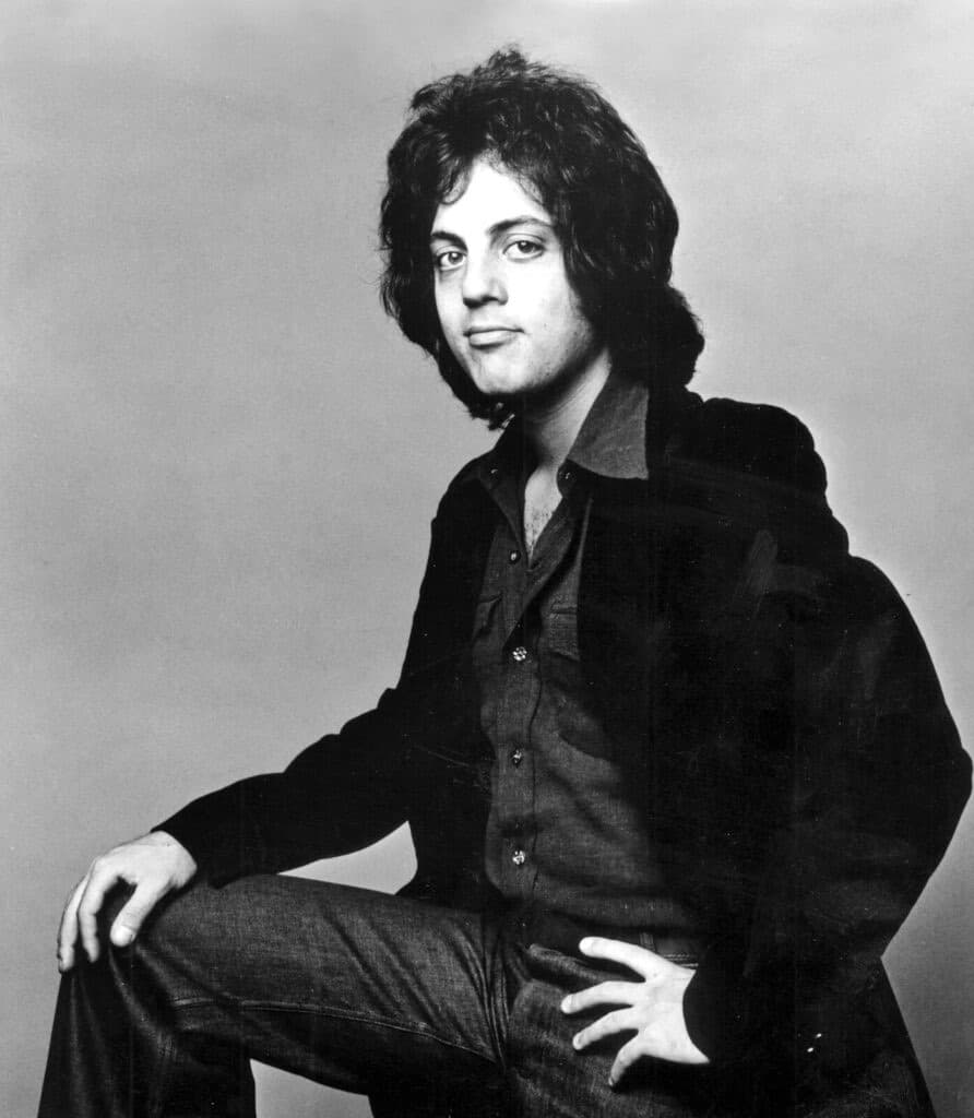 Billy Joel in 1973 sitting with one knee raised, in a dark shirt and jacket