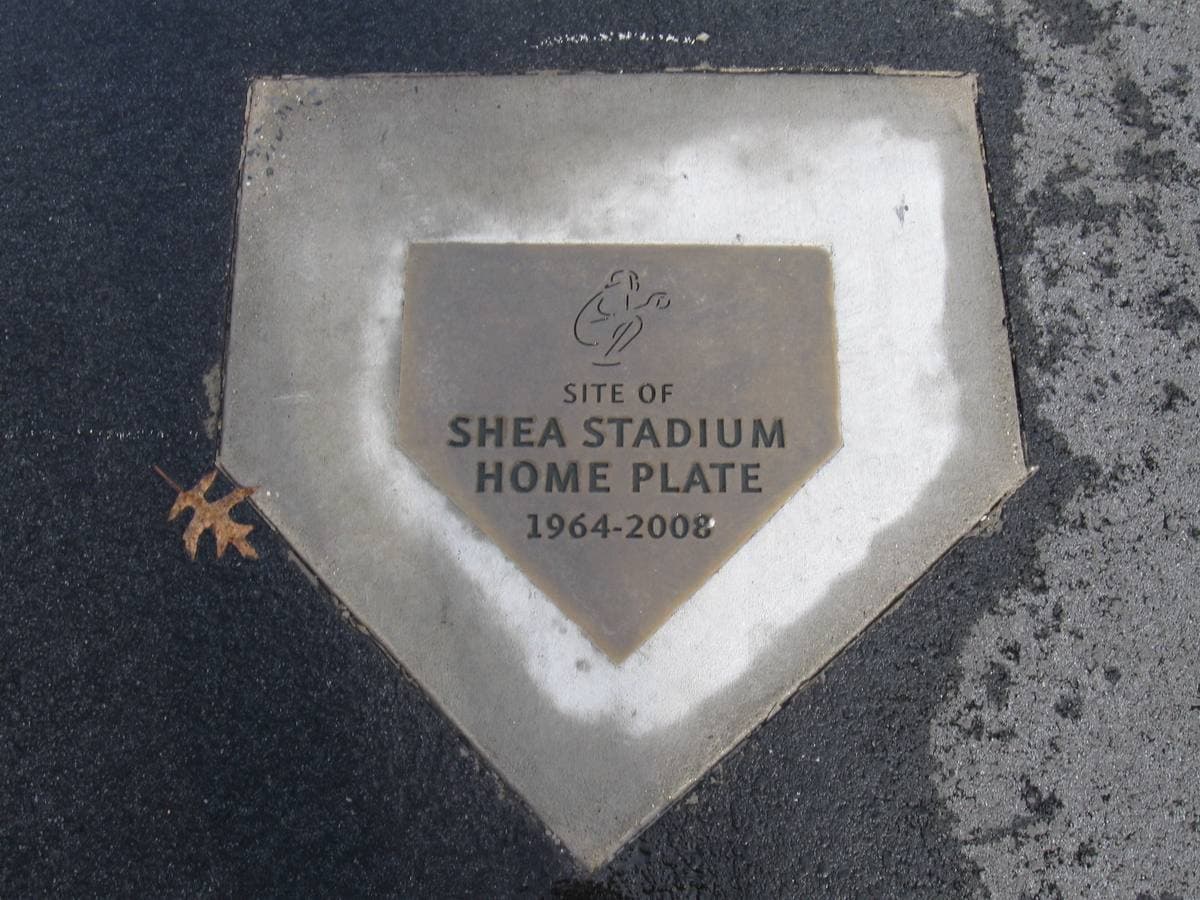 A bronze home-plate shaped plaque reading Site of Shea Stadium Home Plate 1964–2008, set in asphalt