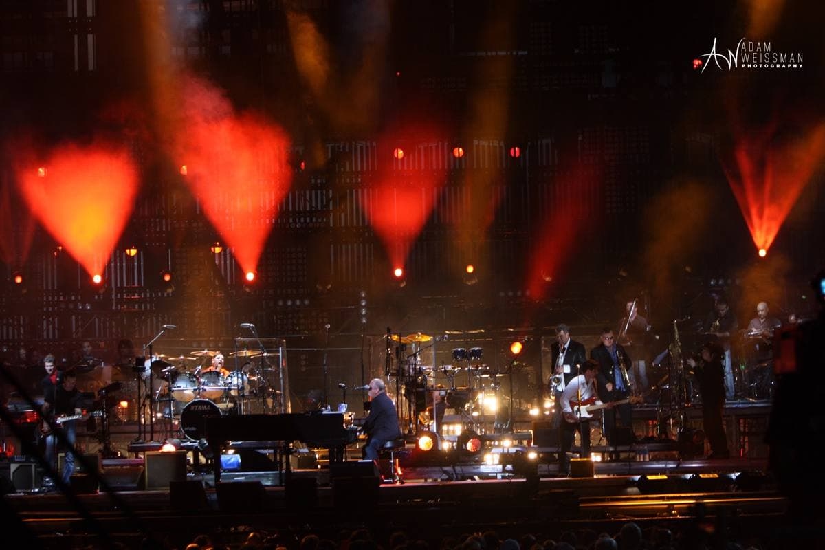 A stadium stage lit red and orange, with a piano and band far below the lights