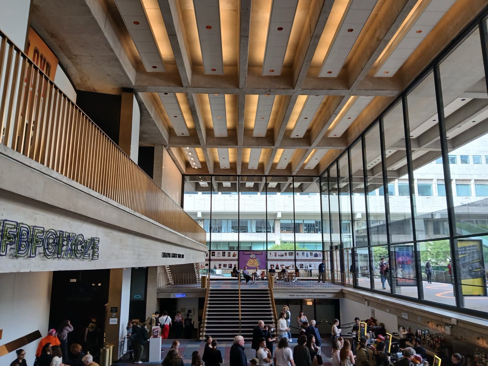 A glass-walled lobby under a timber-ribbed ceiling, people gathering by a wide staircase