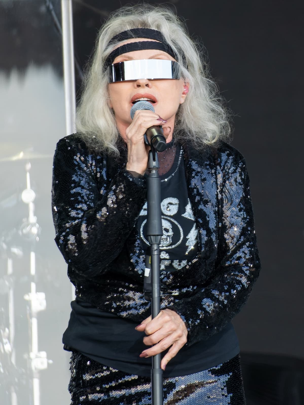 Debbie Harry with silver hair and a mirrored visor singing into a microphone in a sequinned jacket