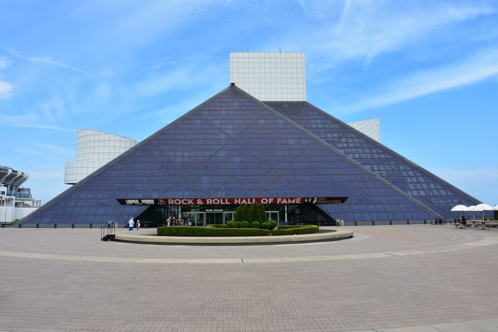 A glass pyramid with a white tower behind it under a blue sky, on a paved plaza