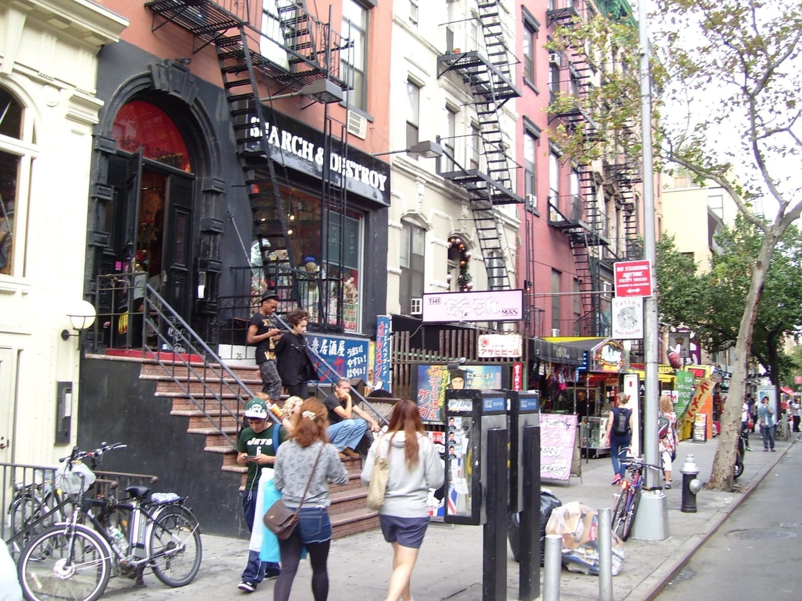 A busy East Village block of brownstones with fire escapes, steps up to shopfronts and people on the pavement