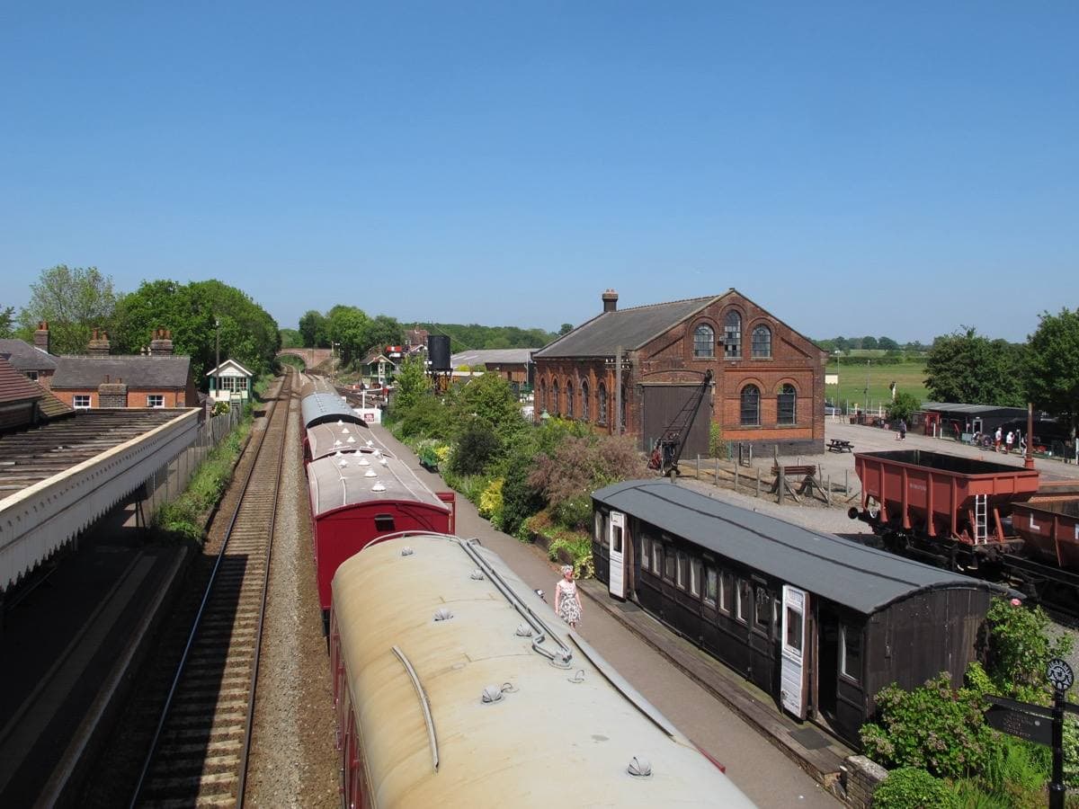 Old railway carriages in a siding beside a platform, with a large Victorian brick goods shed and green fields behind, seen from above