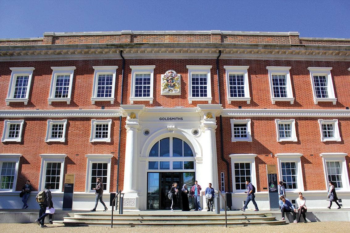 The red-brick front of Goldsmiths' main building with tall white sash windows, a stone arched entrance and students on the steps