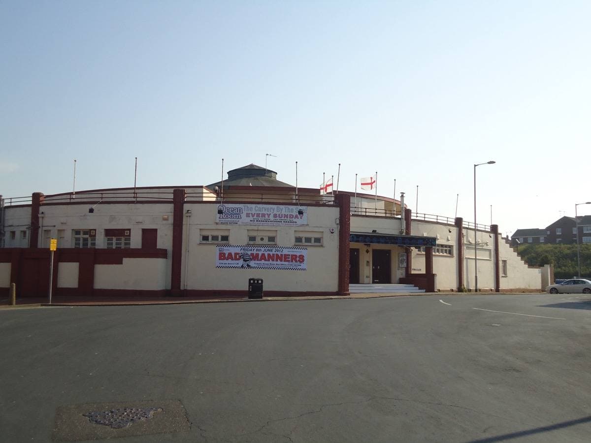 A low white seaside dance hall with red trim and flags on the roof, across an empty car park under a clear sky