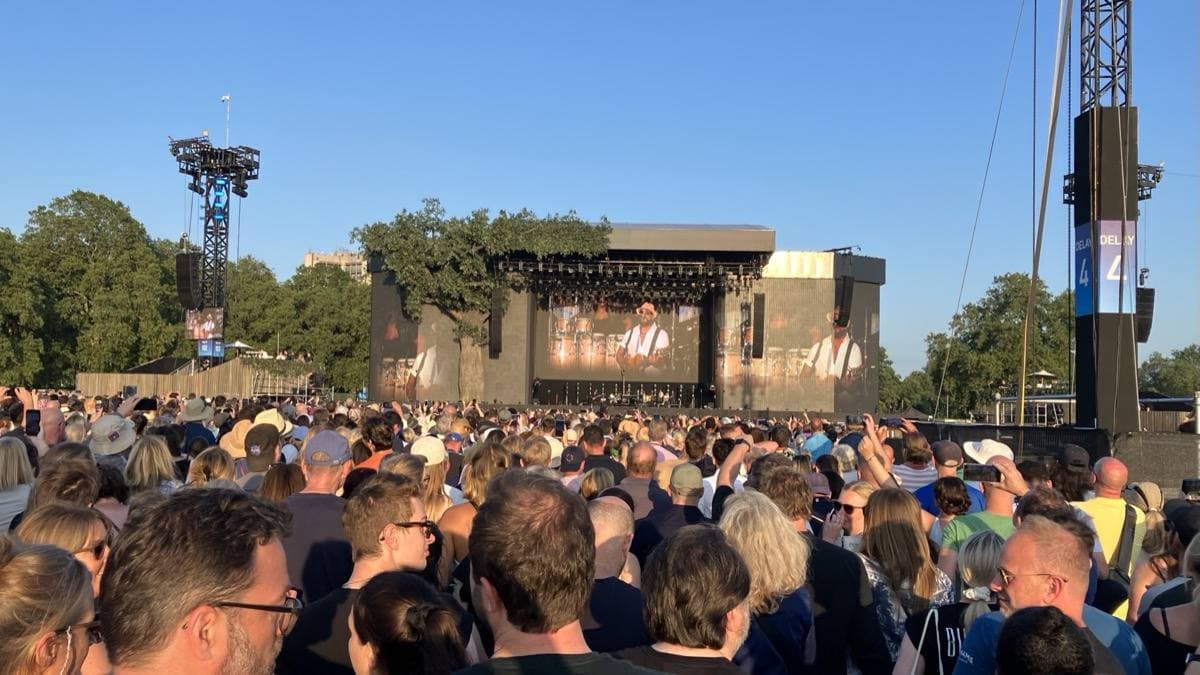 A dense crowd on the grass of Hyde Park facing a large outdoor stage between lighting towers, trees behind, in evening sun