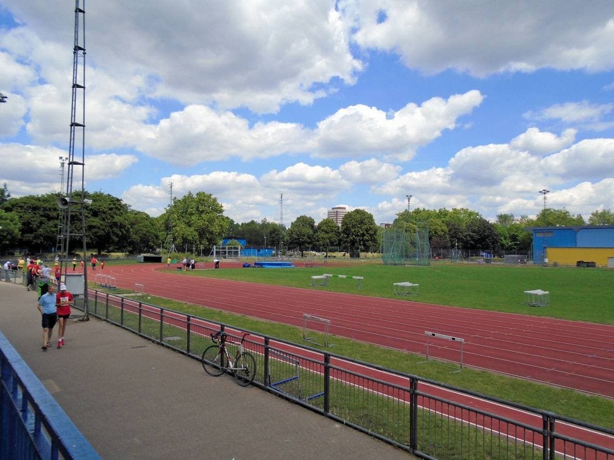 A red running track and green infield under a big summer sky, a blue and yellow stand on the far side and a tower block behind